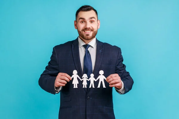 Portrait of happy businessman in official style suit holding paper chain people in hands, concept of happy family, parenthood, childhood. Indoor studio shot isolated on blue background.