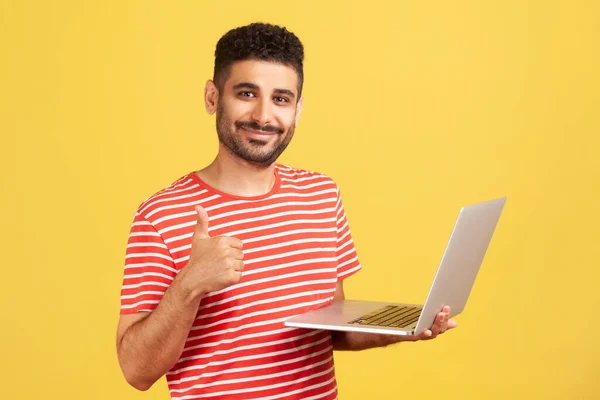 一个满脸笑容的自由职业者 留着胡子 穿着条纹T恤 竖起大拇指 拿着笔记本电脑 对远程办公感到满意 他喜欢他的工作 在黄色背景下隔离的室内拍摄 — 图库照片