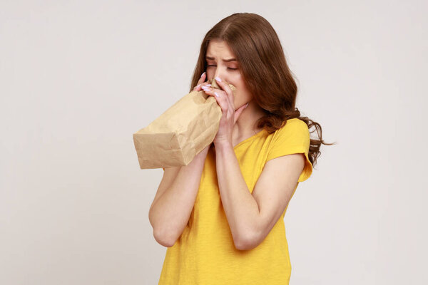 Portrait of nervous teenager girl feeling sick, breathing into paper bag to improve well-being, overcoming stress at work, wearing yellow T-shirt. Indoor studio shot isolated on gray background.