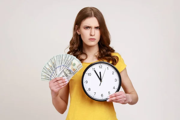 金を稼ぐ時間だ 黄色のTシャツに厳格なティーンエイジャーの少女は カメラの深刻な顔の表情を見て 大きな時計やドル紙幣を保持する 灰色の背景に隔離された屋内スタジオショット — ストック写真