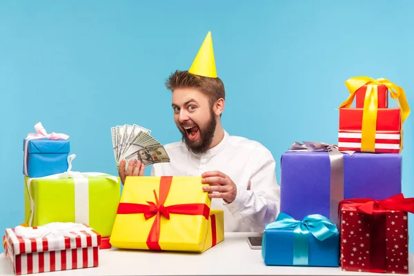 多くのギフトボックスの1つを開き ドルの現金の多くを示すパーティーキャップで幸せな興奮した男は 寛大な現金誕生日プレゼント ボーナスに満足している 青い背景に隔離された屋内スタジオショット — ストック写真