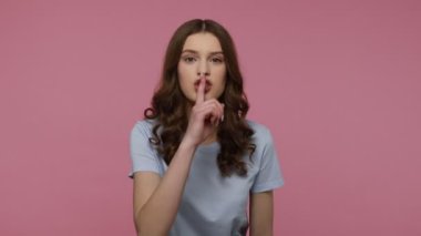 Sus, konuşma! Sıradan tarzda tişörtlü, parmakları dudağında sessiz jestler yapan sessiz kalmak isteyen, ciddi bir ifadeyle kameraya bakan bir kız. Pembe arka planda kapalı stüdyo çekimleri.