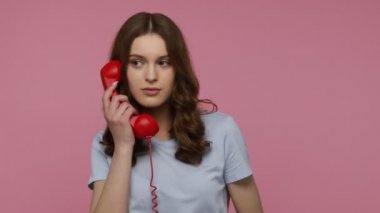 Siyah dalgalı saçlı, sıradan tişörtlü, telefonla konuşan, telefonla konuşan ve müşterilerle iletişim kuran neşeli, arkadaş canlısı genç kız. Pembe arka planda kapalı stüdyo çekimleri.