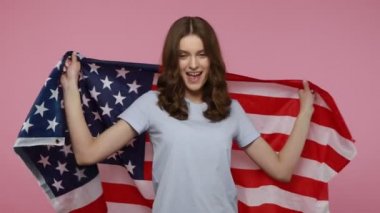 Hoş görünümlü bir kız Amerikan bayrağı sallıyor ve neşeyle gülüyor, Bağımsızlık Günü 'nü kutluyor, mutlu bir ifadeyle kameraya bakıyor. Pembe arka planda kapalı stüdyo çekimleri.