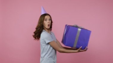 Mavi tişörtlü tatmin olmuş genç kız, neşeli ifadeli büyük hediye kutusunu kusuyor. Meraklı kadın doğum günü hediyesini açıyor. Pembe arka planda kapalı stüdyo çekimleri.