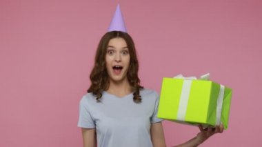 Hediye kutusunu tutan, neşeyle dans eden ve konfeti üfleyen genç, şaşkın ve güzel bir kadın uzun zamandır beklenen hediyeden memnun görünüyor. Pembe arka planda kapalı stüdyo çekimleri