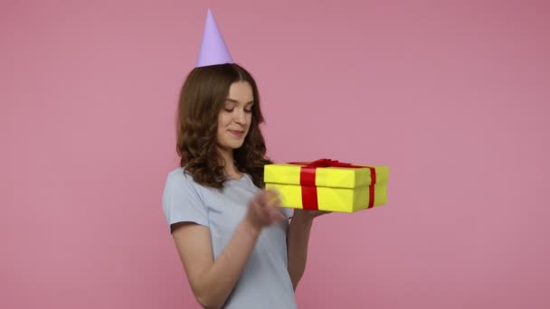Tシャツの愛らしい女の子は ギフトを包み 箱を開けて カメラの喜びを見て 誕生日プレゼントに満足している大きな驚きを表現しています 屋内スタジオはピンクの背景に隔離されたショット — ストック動画