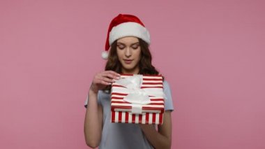 Genç yaşta çekici bir kadın hediye kutusunu açıyor ve sevinçle gülümsüyor, güzel bir Noel hediyesi ile tatmin oluyor, beklenmedik bir doğum günü sürprizi. Pembe arka planda kapalı stüdyo çekimleri.
