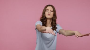 Karmaşık, mavi tişörtlü, gözleri kapalı, görünmez engellere dokunan, karanlıkta yolu kontrol eden çekici, genç bir kadın. Pembe arka planda kapalı stüdyo çekimleri.