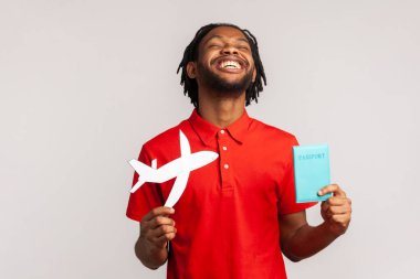 Rastgele giyinen, kırmızı tişörtlü, elinde pasaport ve kağıt uçak tutan, neşeli bir seyahat turu yapan son derece mutlu bir adam. Gri arka planda izole edilmiş kapalı stüdyo görüntüleri.