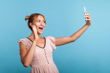 Kıllı kalçalı mutlu bir kadının selfie çekip el sallaması, takipçilere merhaba işareti yapması, video görüşmesiyle iletişim kurması. Mavi arka planda kapalı stüdyo çekimi.