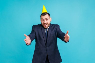 Kollarıma gel! Resmi tarz takım elbiseli ve parti konisi olan arkadaş canlısı bir adam uzanmış elleriyle kucaklaşarak kucaklaşmaya davet ediyor. Mavi arka planda kapalı stüdyo çekimi.