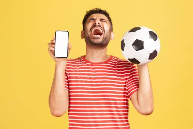 Son derece mutlu, çizgili tişörtlü, sakallı, beyaz akıllı telefon ve futbol topuyla gülen, zaferi kutlayan son derece mutlu bir adam. Sarı arka planda kapalı stüdyo çekimleri