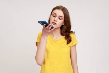 Yenilikçi teknolojiler. Kahverengi dalgalı saçlı genç ve çekici bir kadın cep telefonundan ses asistanı istiyor, görev veriyor, mesaj kaydediyor. Gri arka planda izole edilmiş kapalı stüdyo görüntüleri.