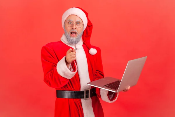 Zszokowany Staruszek Szarą Brodą Kostiumie Świętego Mikołaja Trzymający Laptopa Rękach — Zdjęcie stockowe