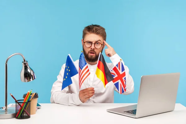 Smart serious man teacher in eyeglasses holding flags of different countries pointing finger at head, suggesting to learn foreign language with him. Indoor studio shot isolated on blue background