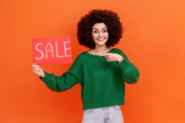 緑のカジュアルなスタイルのセーターを着てアフロの髪型と幸せな笑顔の女性が販売の碑文 巨大な割引でカードを保持立っている オレンジの背景に隔離された屋内スタジオショット — ストック写真