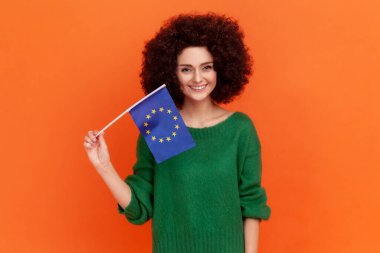 Avrupa bayrağı, Avrupa Birliği birliği ve cemaatin sembolü olarak elinde yeşil günlük süveter giyen Afro saçlı gülümseyen kadın. Turuncu arka planda kapalı stüdyo çekimleri.