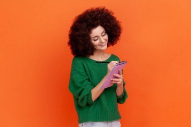 Positive smiling woman with curly hair wearing green casual style sweater writing down in paper notebook, making to do list, having good mood. Indoor studio shot isolated on orange background. clipart