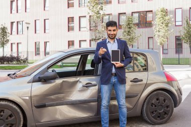 Ceketli ve kot pantolonlu esmer sakallı bir kaza müfettişi boş ekranlı tableti ve kamerayı göstererek sigorta hizmetini seçiyor. Açık hava çekimi.