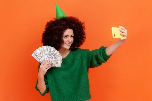 緑色のカジュアルなスタイルのセーターとパーティーコーンを着て巻き毛を持つ笑顔の女性の肖像画は ドル紙幣のファンと自撮りします オレンジの背景に隔離された屋内スタジオショット — ストック写真