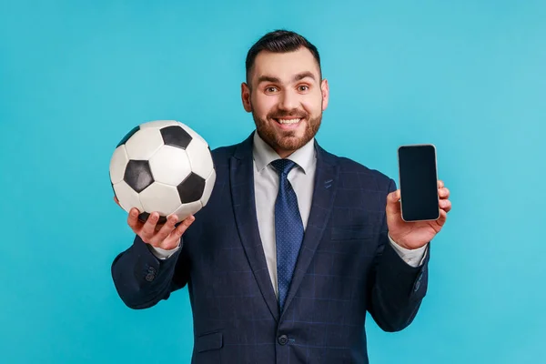 スマートフォンやサッカーボールを空白のディスプレイでスマートフォンを保持公式スタイルのスーツを着て笑顔ビジネスマンは サッカーに賭ける 青い背景に隔離された屋内スタジオショット — ストック写真