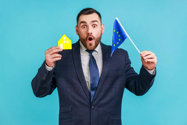 Amazed man with beard wearing official style suit holding European flag and paper house, looking at camera with surprised expression. Indoor studio shot isolated on blue background.