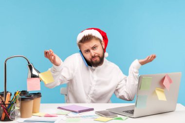 Noel Baba şapkalı kafası karışmış bir adam omuz silkiyor, telefonla konuşuyor, iş yerinde dizüstü bilgisayarla oturuyor, tatillerde ve teslim tarihlerinde çalışıyor. Mavi arka planda kapalı stüdyo çekimi