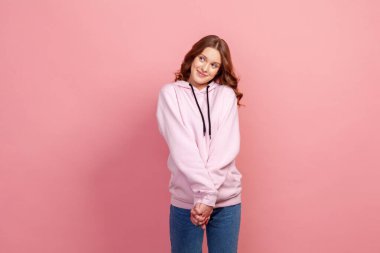 Portrait of adorable dreamy young woman with brown hair in hoodie standing, holding palms together while dreaming, making wish, love. Indoor studio shot isolated on pink background clipart