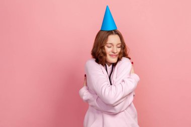 Kafasında parti konisi olan, kendini kucaklayan ve mutlu bir ifadeyle gülümseyen kapüşonlu, kıvırcık saçlı genç kızın portresi. Pembe arka planda kapalı stüdyo çekimleri