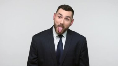 Siyah resmi takım elbiseli komik, pozitif sakallı iş adamı dilini gösteriyor, çocuksu berhavior, şeytani bir ifade sergiliyor. Gri arka planda izole edilmiş kapalı stüdyo görüntüleri.