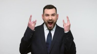 Aşırı mutlu sakallı iş adamının portresi siyah resmi kıyafet içinde heyecanla bağırıyor ve kameraya rock 'n roll hareketi gösteriyor. Gri arka planda izole edilmiş kapalı stüdyo görüntüleri.