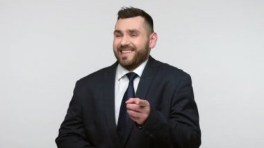 Siyah resmi takım elbiseli mutlu, pozitif sakallı iş adamının portresi kamerayı işaret ediyor, seni seçiyor ve yüksek sesle gülüyor. Gri arka planda izole edilmiş kapalı stüdyo görüntüleri.