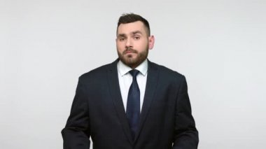 Siyah resmi takım elbiseli, kafası karışmış, sakalsız iş adamı omuz silkiyor, doğru cevabı bilmiyor, şüpheli bir ifade takınıyor. Gri arka planda izole edilmiş kapalı stüdyo görüntüleri.