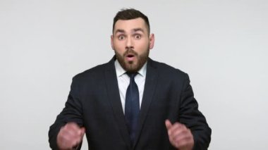 Siyah resmi takım elbiseli aşırı derecede şok olmuş sakallı bir işadamı kameraya büyük gözlü ve ağzı açık bakıyor ve hayret verici bir şey görüyor. Gri arka planda izole edilmiş kapalı stüdyo görüntüleri.