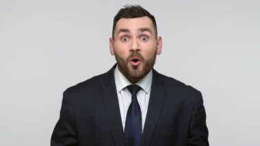 Siyah resmi takım elbiseli genç ve sakallı bir iş adamının portresi ağzını açık tutuyor ve şok edici haberler duyuyor. Gri arka planda izole edilmiş kapalı stüdyo görüntüleri.