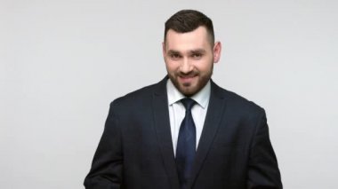 Siyah resmi kıyafetli genç, sakallı iş adamının portresi sinsice gülümsüyor, kurnaz bir planı var, kameraya bakıyor, sinsi bir ifade. Gri arka planda izole edilmiş kapalı stüdyo görüntüleri.