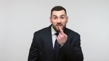 Siyah resmi takım elbiseli agresif sakallı iş adamı yalan jestler yapıp kameraya öfkeyle bakıyor. Gri arka planda izole edilmiş kapalı stüdyo görüntüleri.