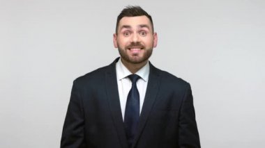 Siyah resmi takım elbiseli iyimser sakallı iş adamı kendini göstererek kameraya dişlek bir gülümsemeyle bakıyor, son derece memnun. Gri arka planda izole edilmiş kapalı stüdyo görüntüleri.