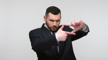 Siyah resmi takım elbiseli meraklı sakallı işadamı el yapımı fotoğraf çerçevesine bakıyor, ilgiyle uzaktan bakıyor. Gri arka planda izole edilmiş kapalı stüdyo görüntüleri.