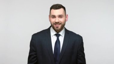 Siyah takım elbiseli, iyi görünümlü, pozitif sakallı iş adamı bana jest diyor, mutlu bir ifadeyle kameraya bakıyor. Gri arka planda izole edilmiş kapalı stüdyo görüntüleri.