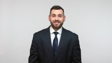 Siyah resmi takım elbiseli mutlu sakallı iş adamı geniş açık ellerle seni selamlıyor. Sevinçle bakıyor, dişlek bir gülümsemeyle. Gri arka planda izole edilmiş kapalı stüdyo görüntüleri.