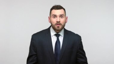 Siyah resmi takım elbiseli, ağzı kapatan, şok edici sırları duyan, büyük gözlerle kameraya bakan hayret verici sakallı iş adamı. Gri arka planda izole edilmiş kapalı stüdyo görüntüleri.