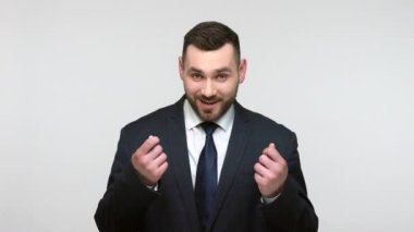 Siyah resmi takım elbiseli sakallı iş adamı para hareketi yapıyor ve başını sallıyor, harçlık istiyor, büyük gelir planlıyor ve gülümsüyor. Gri arka planda izole edilmiş kapalı stüdyo görüntüleri.