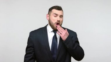 Siyah takım elbiseli yakışıklı bir iş adamı. Elleriyle ağzını kapatıyor. Sıkılmış ya da kayıtsız kalmış, uykusuzluk çekiyor. Gri arka planda izole edilmiş kapalı stüdyo görüntüleri.