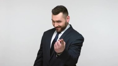 Siyah resmi takım elbiseli genç, sakallı bir iş adamı, jest yapıyor, buluşmaya davet ediyor, oynak cilveli görünüyor. Gri arka planda izole edilmiş kapalı stüdyo görüntüleri.