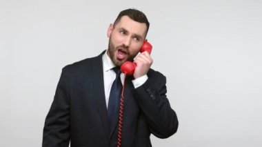 Siyah resmi takım elbiseli hoş görünümlü genç, sakallı iş adamı müşteriyle konuşuyor, kırmızı sabit telefon kullanıyor, gülümsüyor. Gri arka planda izole edilmiş kapalı stüdyo görüntüleri.