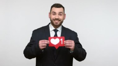 Siyah resmi kıyafet giymiş neşeli sakallı iş adamının portresi sosyal medyayı gösteriyor. Simge, kalp tuşu, en iyi blog içeriğini seç. Gri arka planda izole edilmiş kapalı stüdyo görüntüleri.