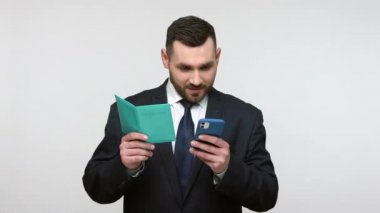 Siyah resmi kıyafet giymiş, elinde pasaport ve akıllı telefon olan çekici sakallı iş adamının portresi, veri girişi, rezervasyon turu. Gri arka planda izole edilmiş kapalı stüdyo görüntüleri.