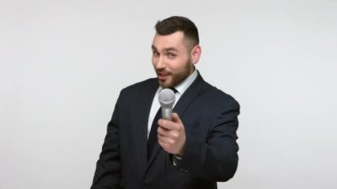 Siyah resmi kıyafetli, sakallı bir adamın portresi mikrofon sunuyor, gazetecilerle röportaj yapıyor, questinlere soruyor, kameraya bakıyor. Gri arka planda izole edilmiş kapalı stüdyo görüntüleri.